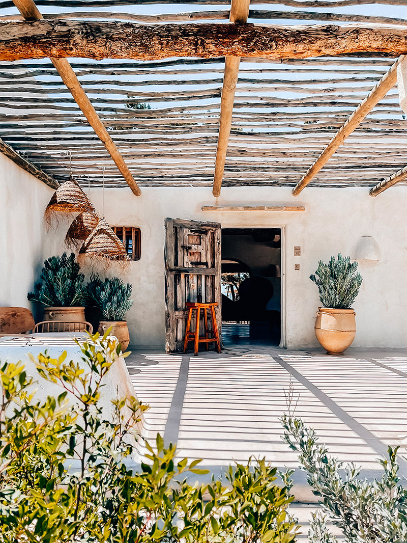 In deze fijne Ibiza tuin is er gekozen voor een luchtige natuurlijke overkapping van houten pilaren en takken, waarmee een fijn schaduwplekje is gecreëerd voor de eethoek.