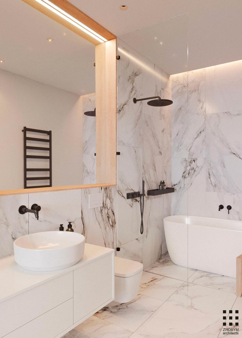 Zrobym Architects heeft in deze fijne luxe badkamer gekozen voor witte marmeren tegels en zwarte accenten. In de luxe douche vind je naast een zwarte douchekop en kraan, ook een bijpassende wandplank.