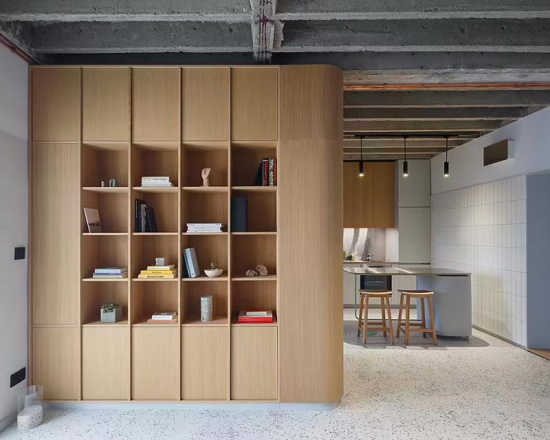 De op maat gemaakte boekenkast met zowel dichte kasten als open vakken wordt als een soort scheidingswand kast gebruikt tussen de woonkamer en half open keuken.
